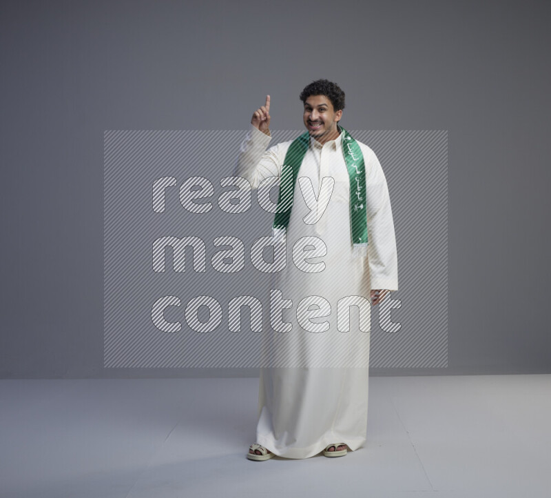 A Saudi man standing wearing thob and Saudi flag scarf on gray background