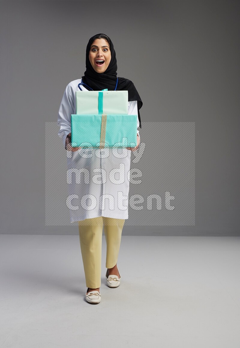 Saudi woman wearing lab coat with stethoscope standing holding gift box on Gray background