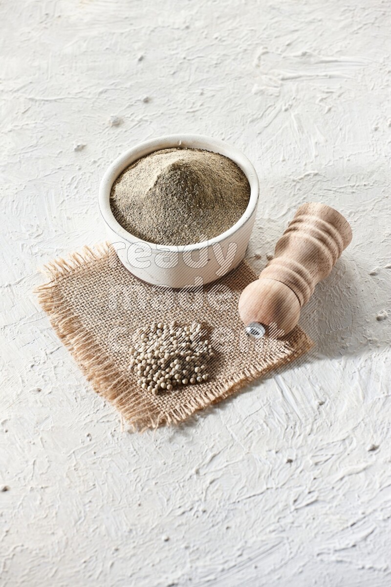White pottery bowl full of white pepper powder set on a burlap piece of fabric with pepper beads and wooden pepper grinder on textured white flooring