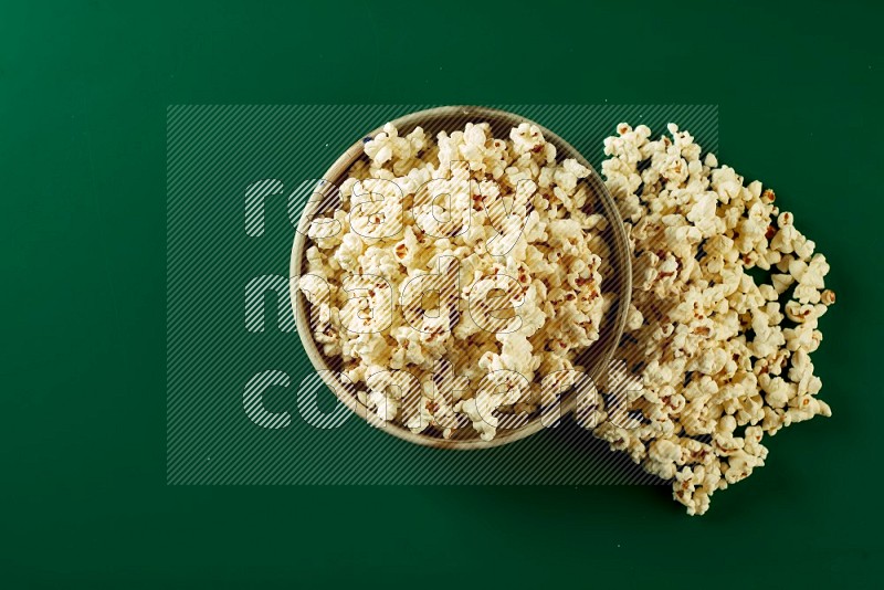 A beige ceramic bowl full of popcorn with popcorn beside it on a green background in a top view shot