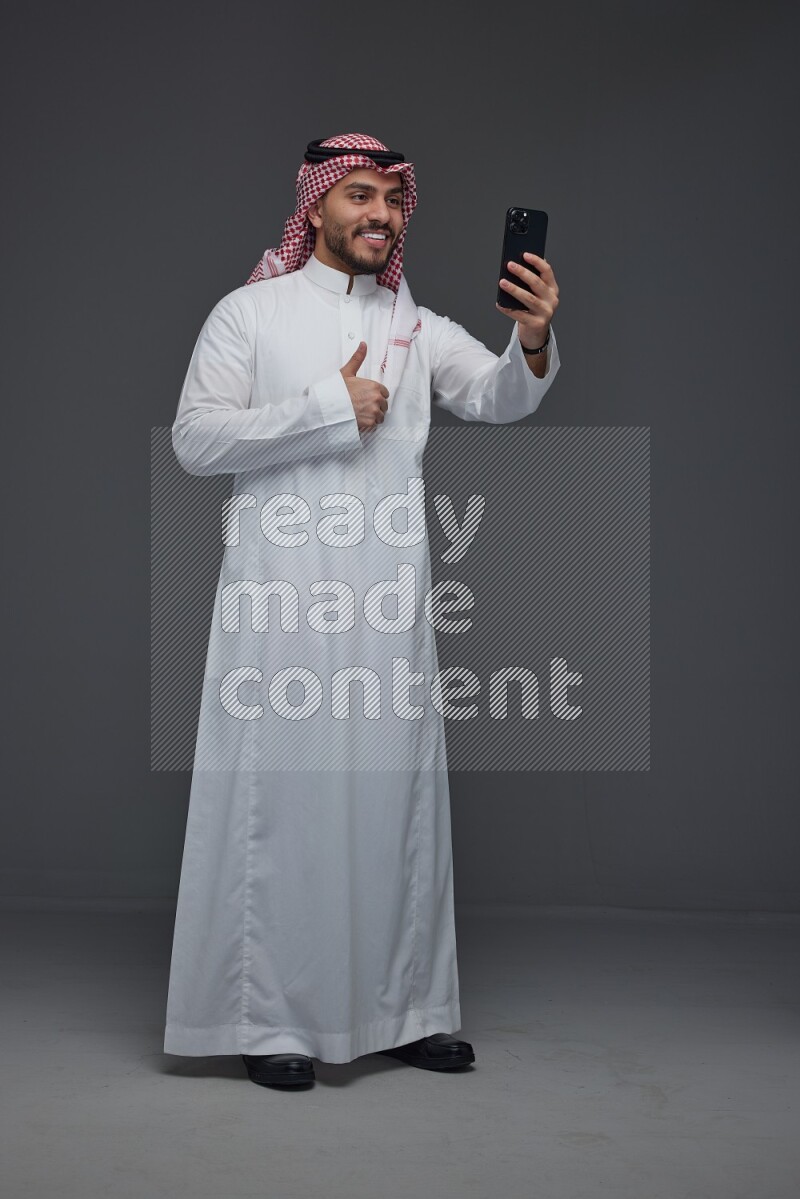 A Saudi man wearing Thobe and Shamgh taking selfie with his phone eye level on a gray background