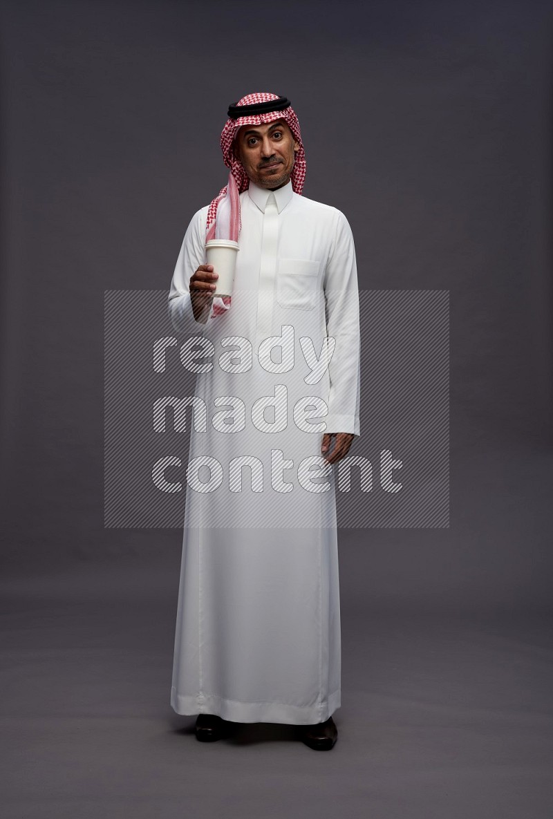 Saudi man wearing thob and shomag standing holding paper cup on gray background