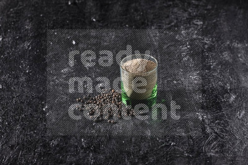 A glass cup full of black pepper powder with beads beside it on a textured black flooring