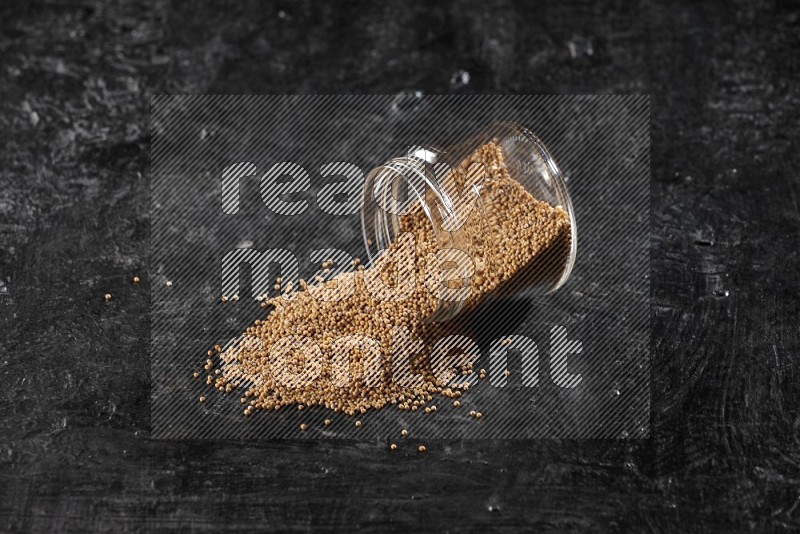 A glass jar full of mustard seeds and jar is flipped and seeds spread out on a textured black flooring