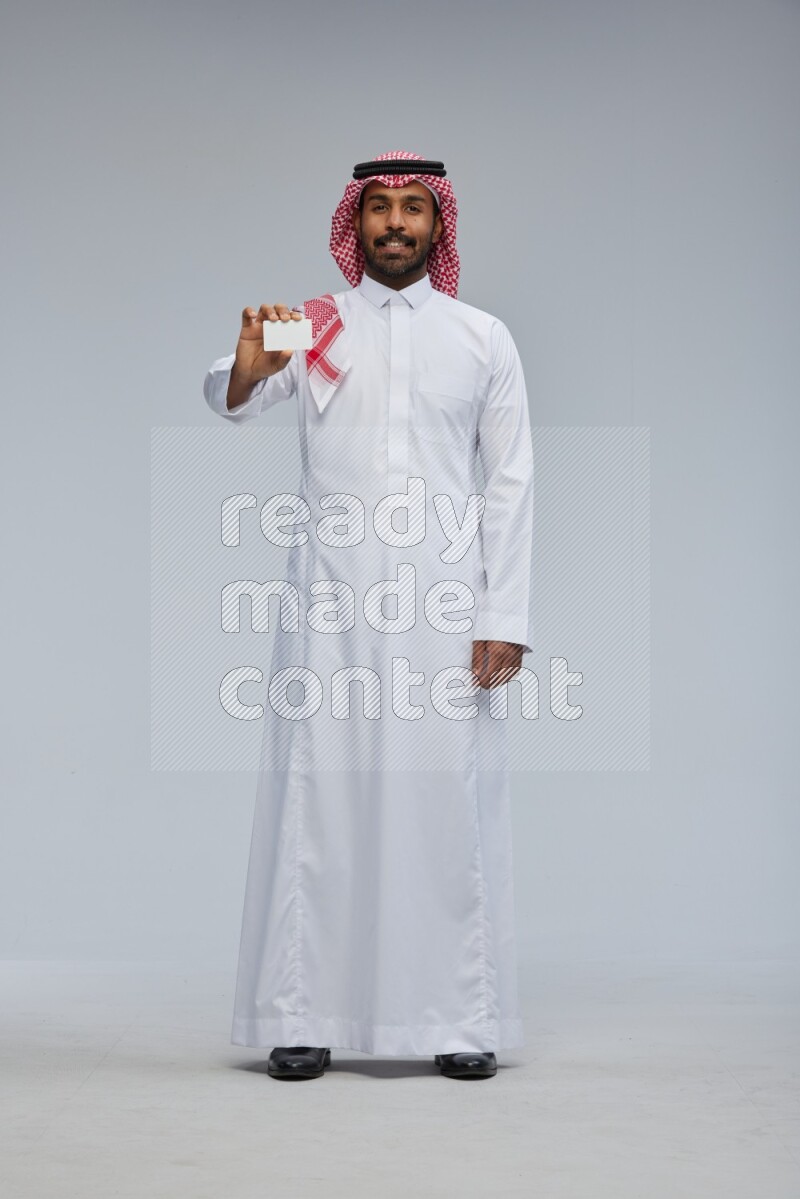 Saudi man wearing thob and shomag standing holding ATM card on gray background