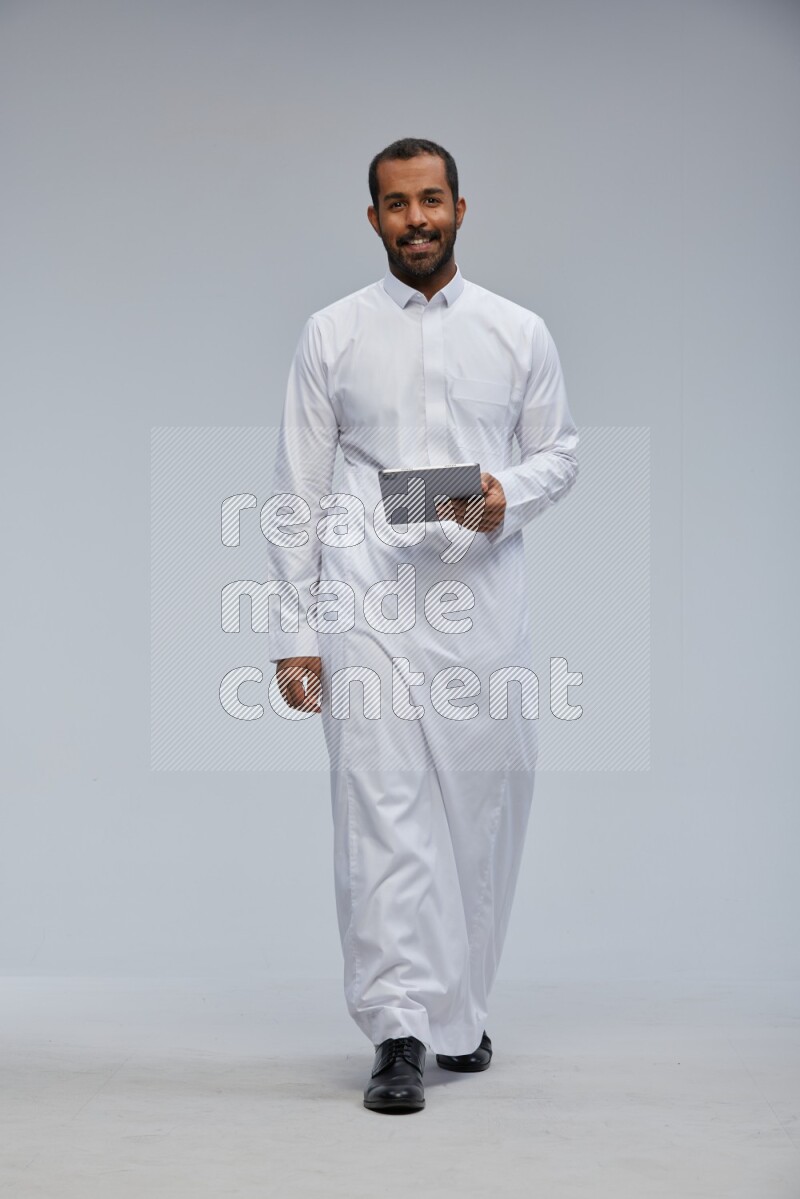 Saudi man wearing Thob standing working on tablet on Gray background