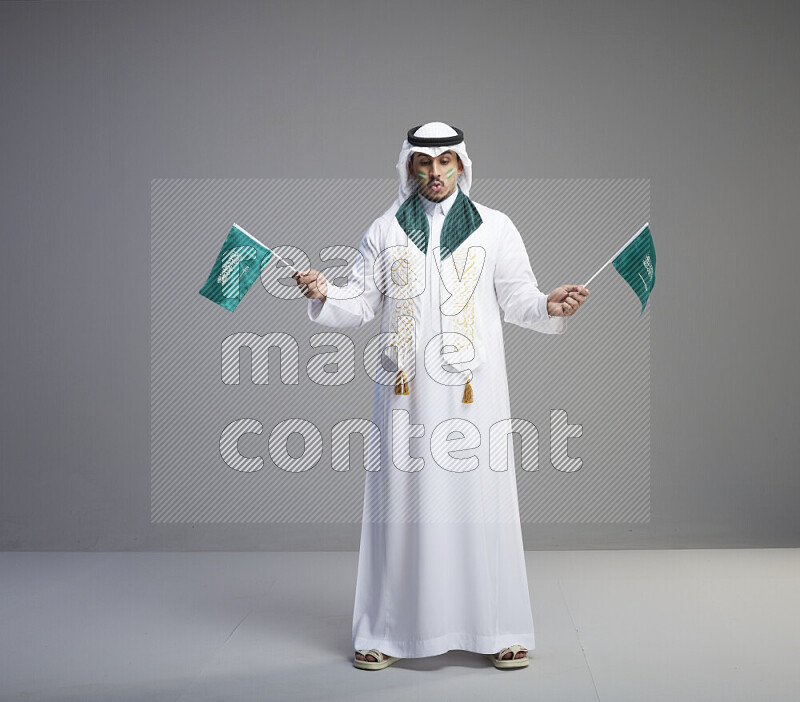 A saudi man standing wearing thob and white shomag with face painting and saudi flag scarf and holding small saudi flag on gray background