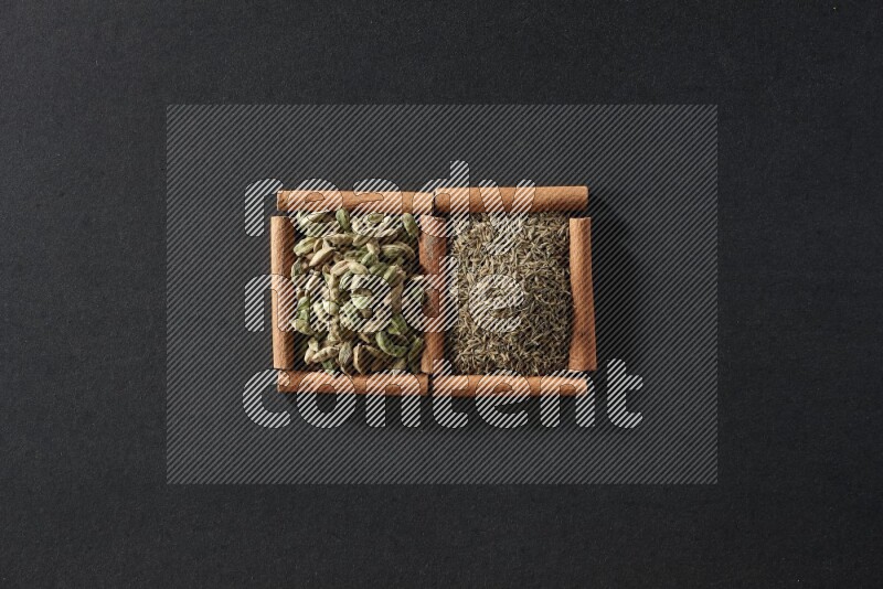 مربعان من أعواد القرفة ممتلئان بحبوب الكمون وحبوب الهيل علي خلفية سوداء