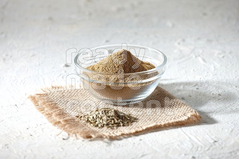 A glass bowl full of cumin powder with some of cumin seeds on burlap piece on a textured white flooring