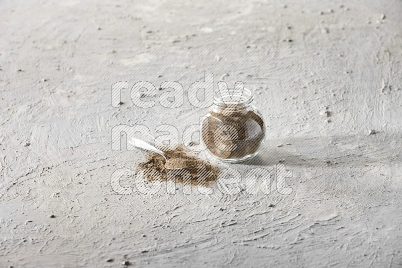 A glass spice jar full of black pepper powder and metal spoon full of it on textured white flooring