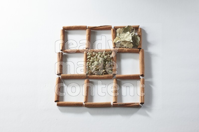 9 مربعات من أعواد القرفة ممتلئة بالهيل في المنتصف محاطة بجوزة الطيب والقرفة وأوراق الغار والقرنفل والكمون والريحان المجفف والزنجبيل المجفف واليانسون النجمي على أرضية بيضاء