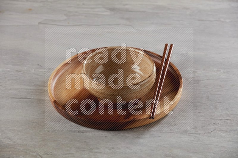 multi color pottery round bowl on top of brown wood round plate and wood chopsticks, on grey textured countertop