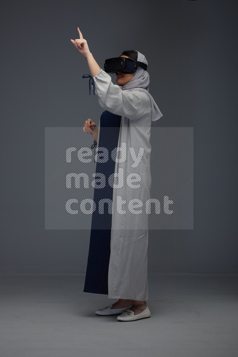 A Saudi woman wearing a light gray Abaya and head scarf standing and wearing VR eye level on a grey background