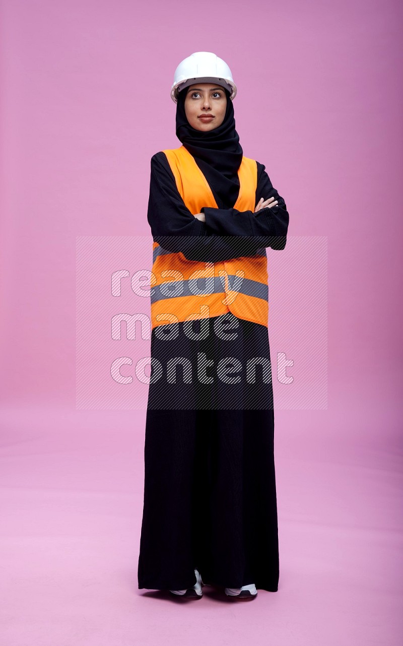 Saudi woman wearing Abaya with engineer vest and helmet standing with crossed arms on pink background