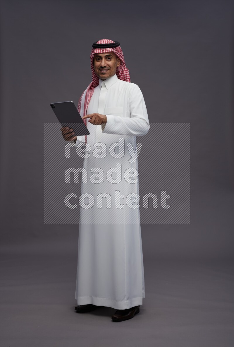 Saudi man wearing thob and shomag standing working on tablet on gray background