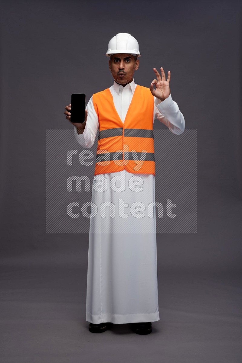 Saudi man wearing thob with engineer vest standing showing phone to camera on gray background