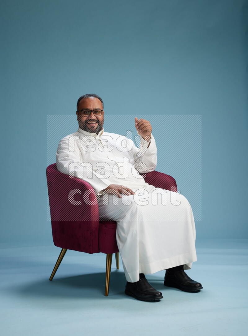Saudi Man without shimag sitting on chair Interacting with the camera on blue background