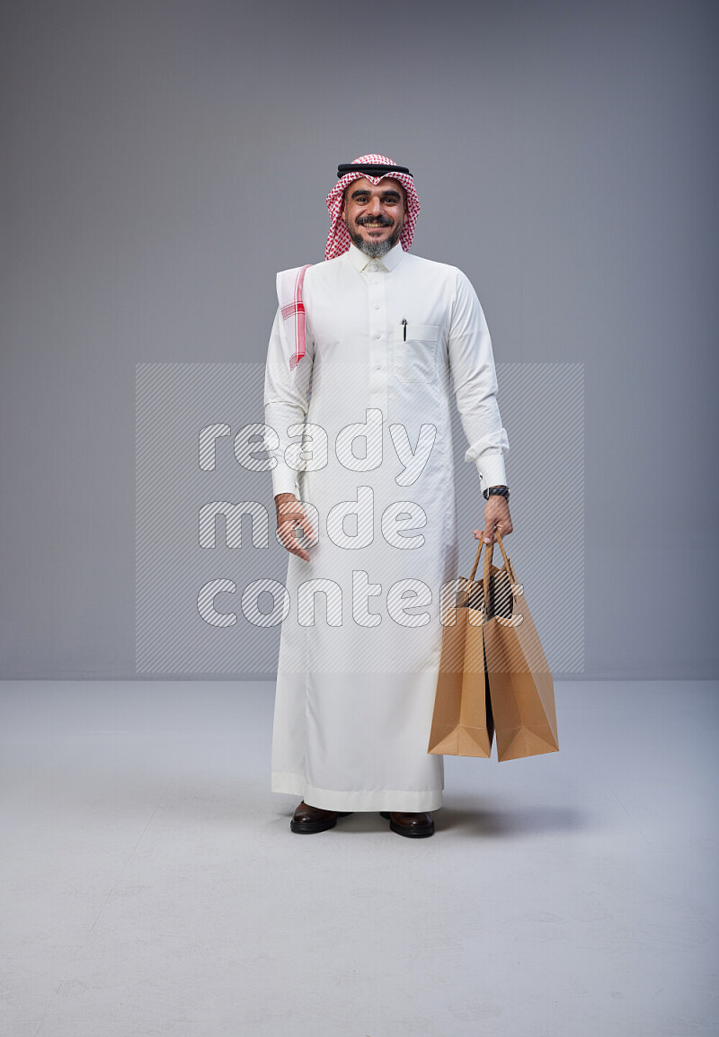 Saudi man Wearing Thob and red Shomag standing holding shopping bag on Gray background