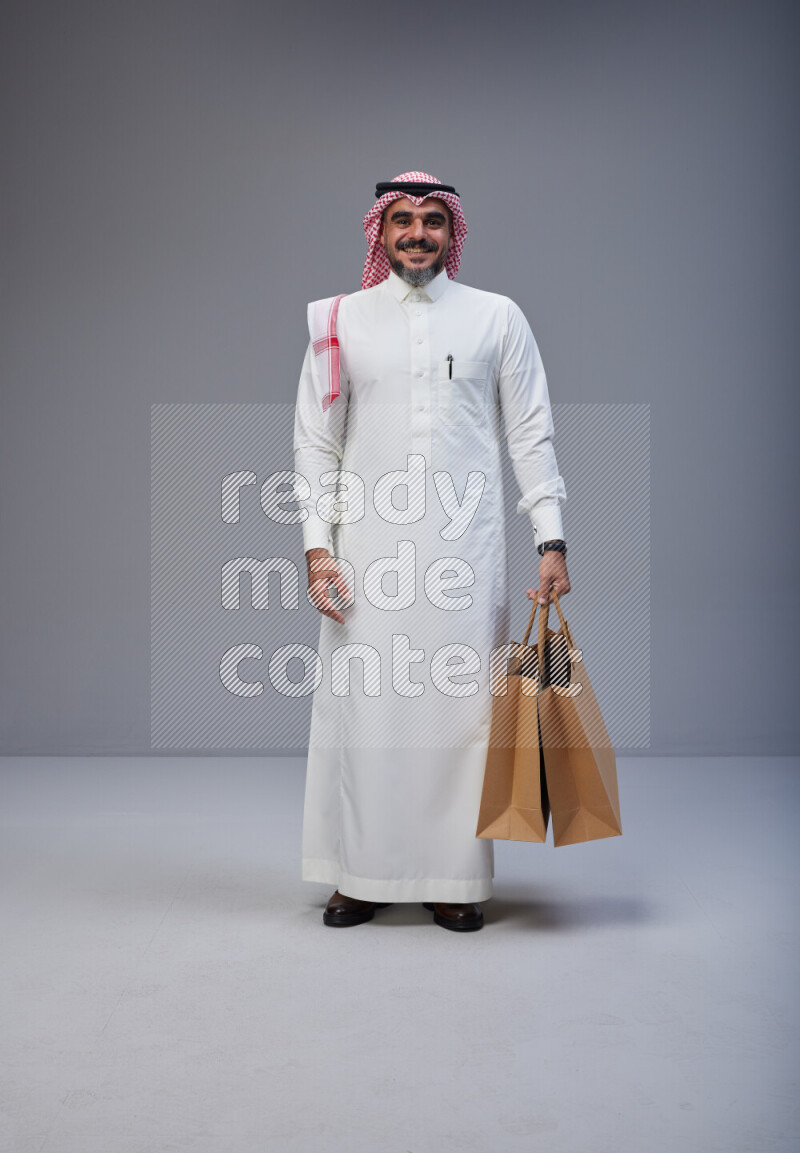 Saudi man Wearing Thob and red Shomag standing holding shopping bag on Gray background