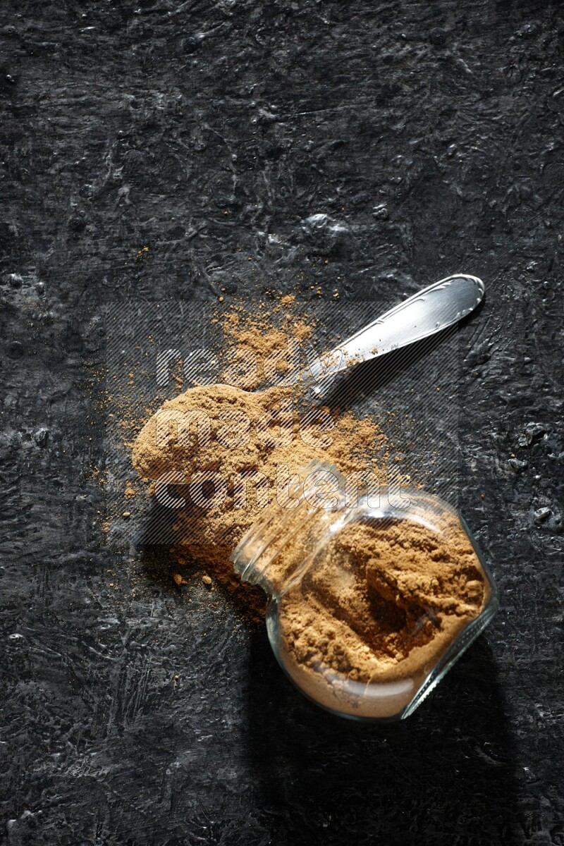 A flipped glass spice jar and a metal spoon full of allspice powder and powder spilled out of it on a textured black flooring