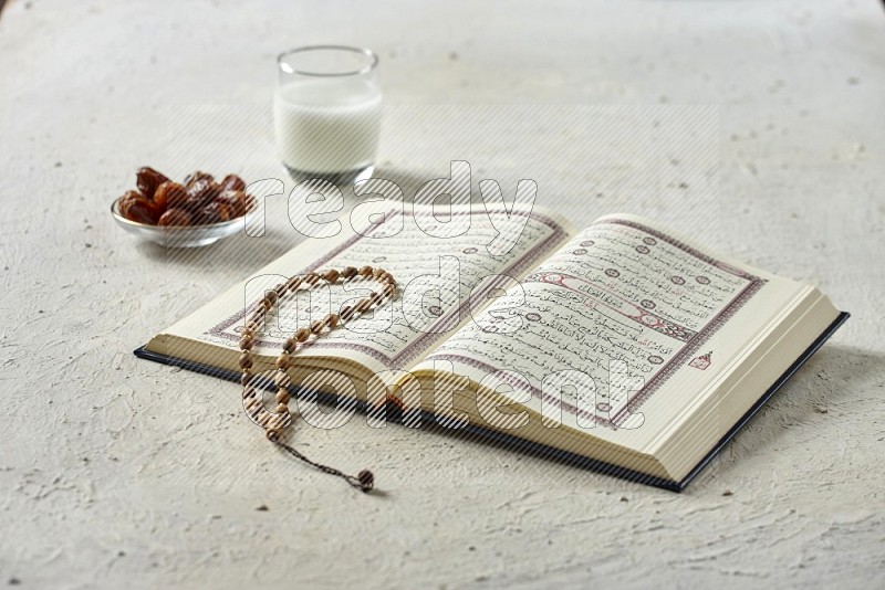 Quran with dates, prayer beads and different drinks all placed on textured white background