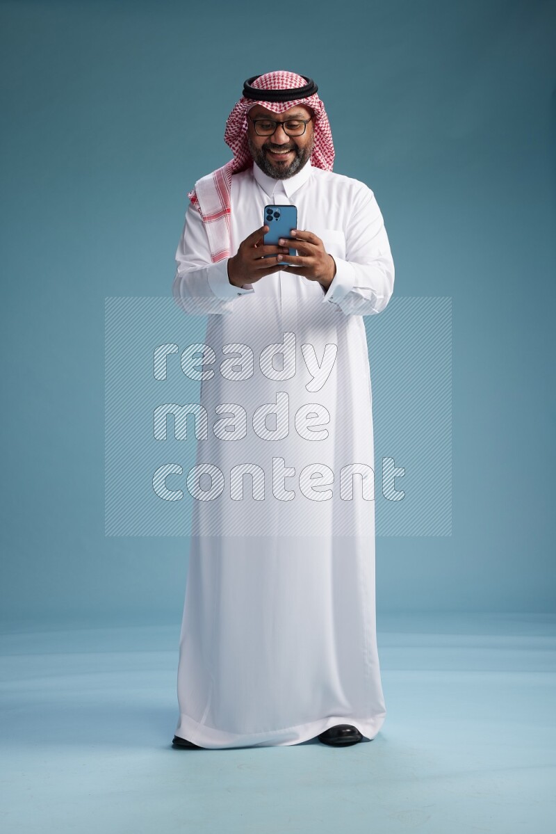 Saudi Man with shimag Standing texting on phone on blue background