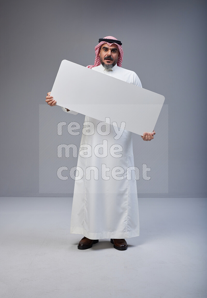 Saudi man Wearing Thob and red Shomag standing holding board on Gray background