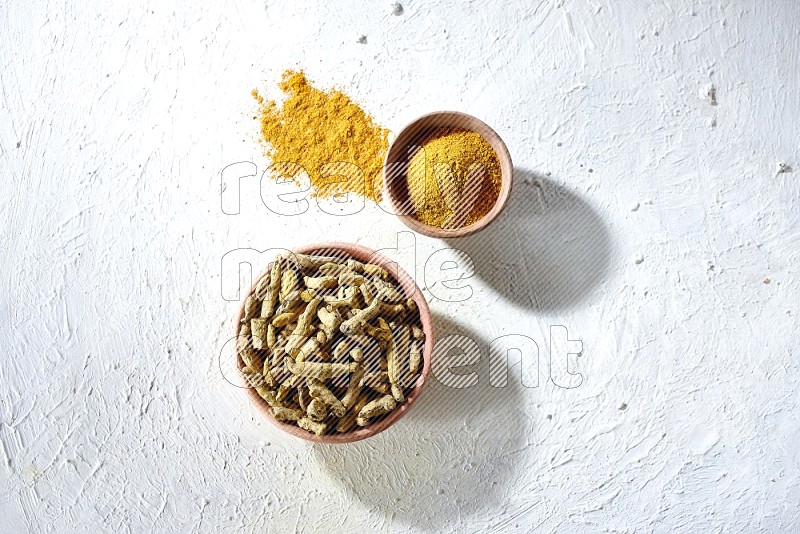 2 wooden bowls, one full of turmeric powder and the other full of dried turmeric whole fingers on a textured white flooring