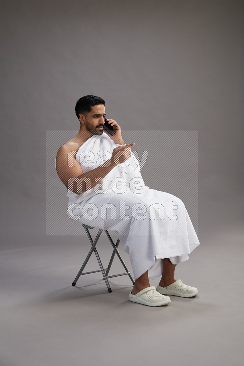 A man wearing Ehram sitting on chair talking on the phone on gray background