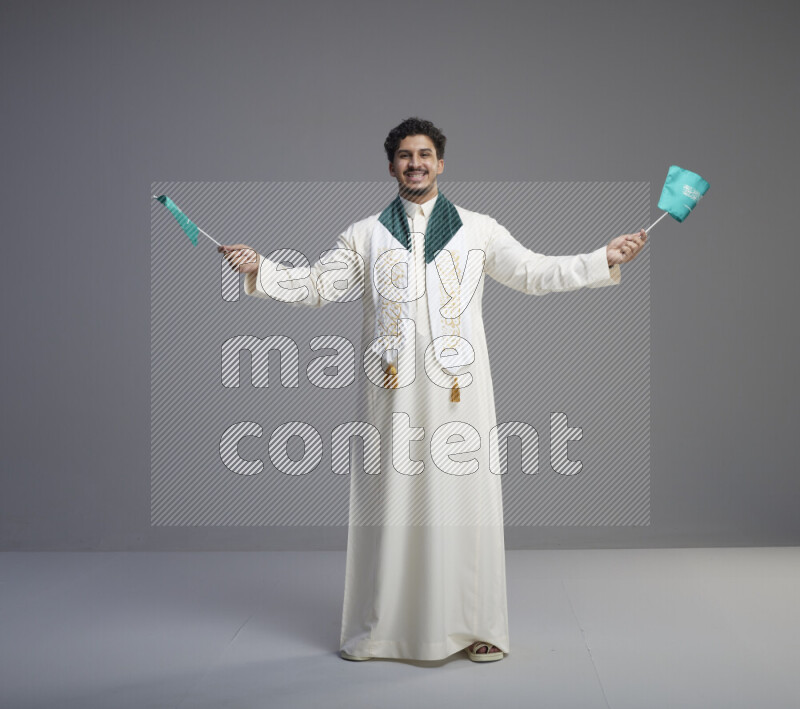 A Saudi man standing wearing thob and saudi flag scarf and holding small saudi flag on gray background