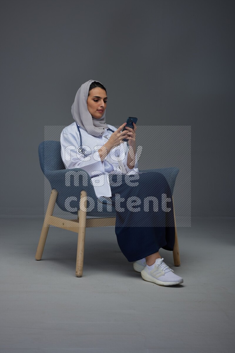 A doctor wearing a light gray head scarf sitting on a dark grey chair and using a laptop while talking in the phone eye level on a grey background