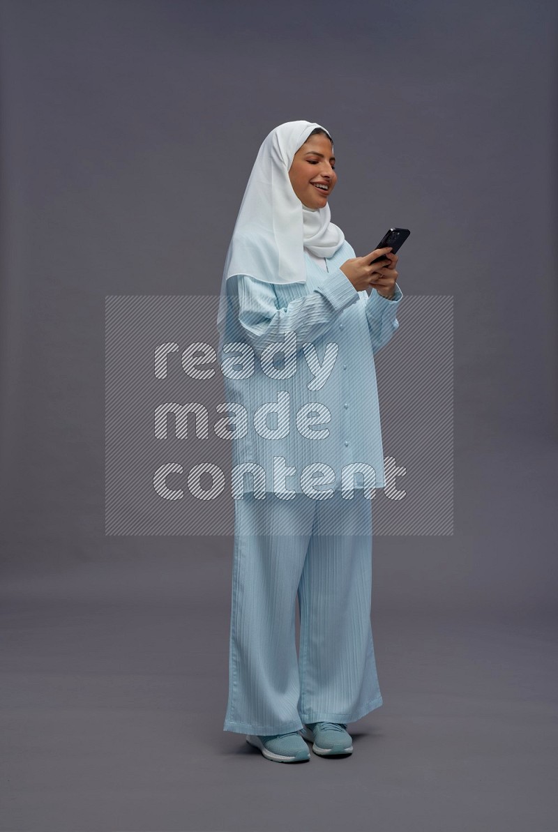 Saudi woman wearing hijab clothes standing texting on phone on gray background