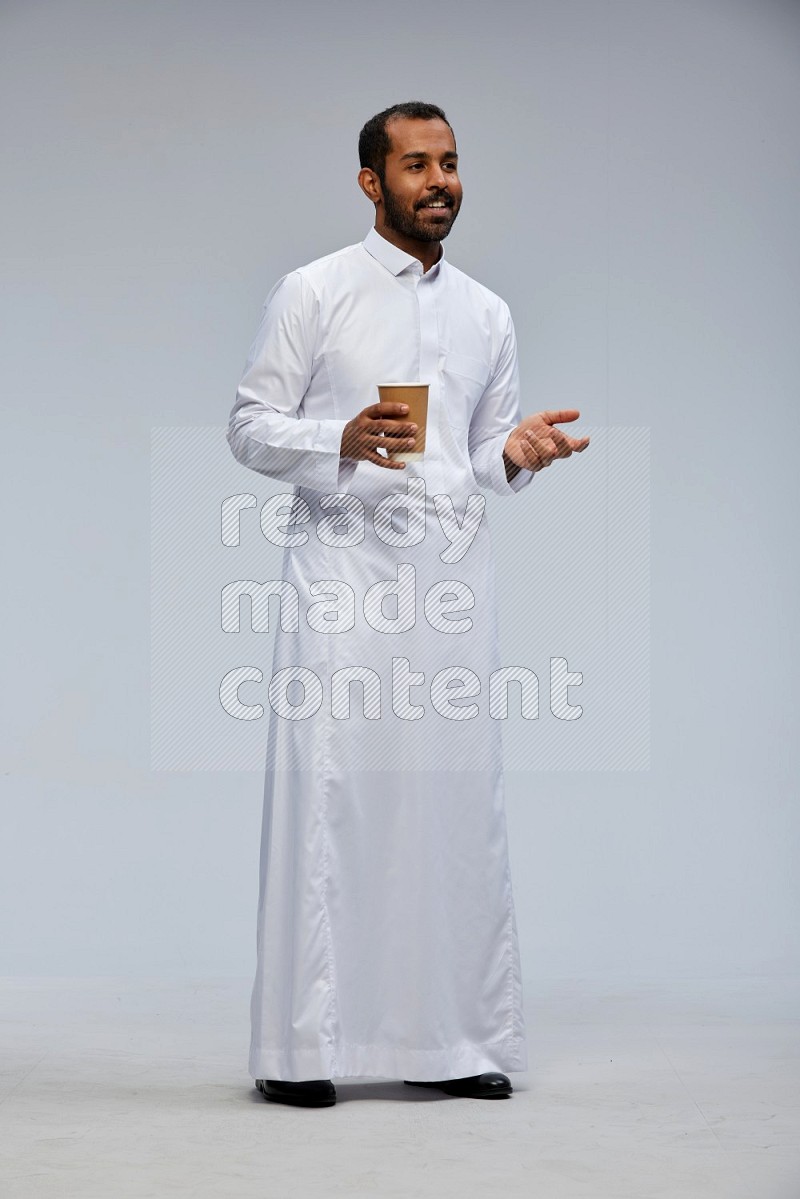 Saudi man wearing thob standing holding paper cup on gray background