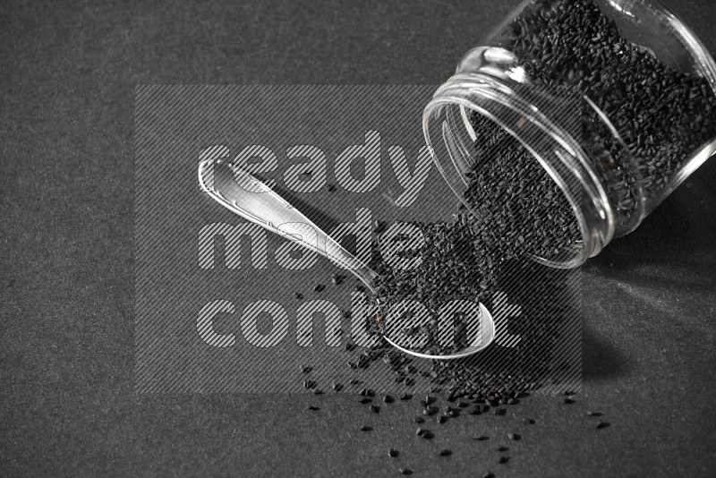 A flipped glass jar full of black seeds and seeds spread out with a metal spoon full of the seeds on a black flooring