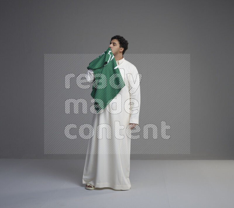 A Saudi man standing wearing thob kissing Saudi flag on gray background