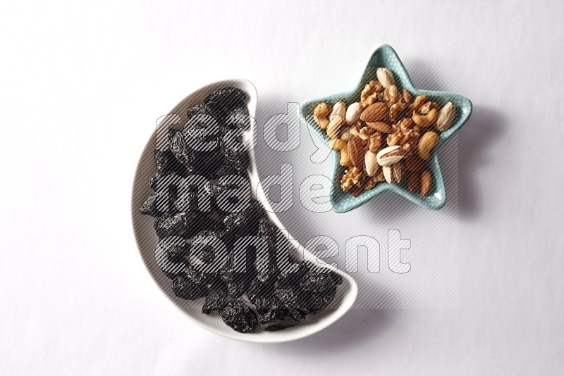 Dried plums in a crescent pottery plate and a star shaped plate with mixed nuts on white background