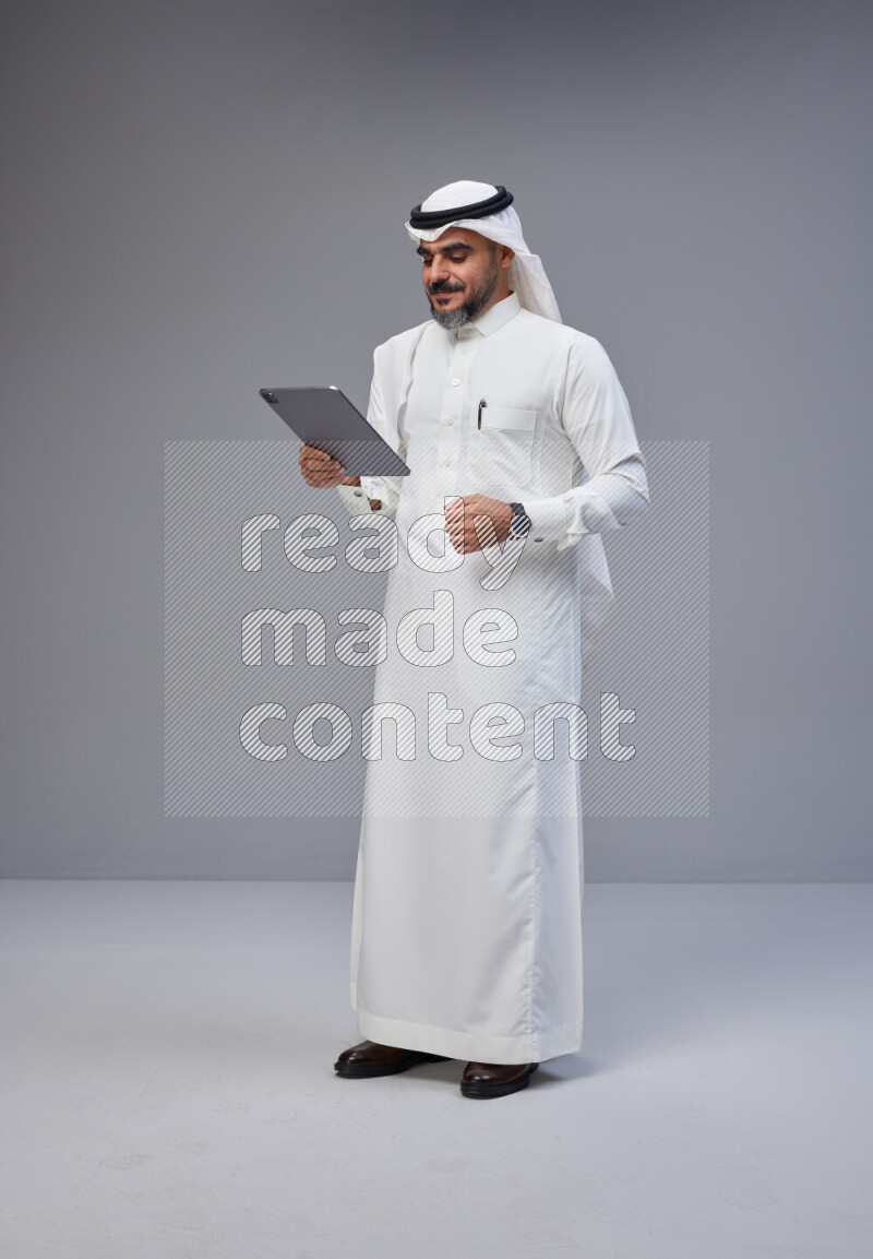 Saudi man Wearing Thob and white Shomag standing working on tablet on Gray background