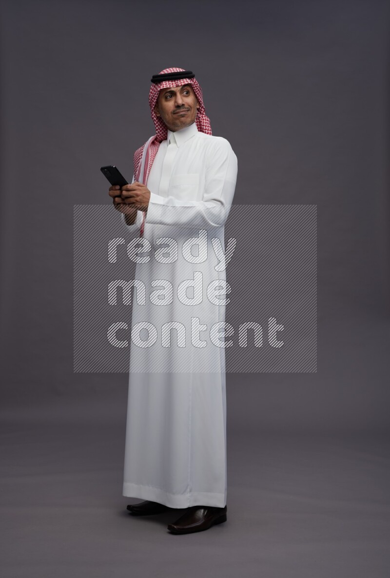 Saudi man wearing thob and shomag standing texting on phone on gray background