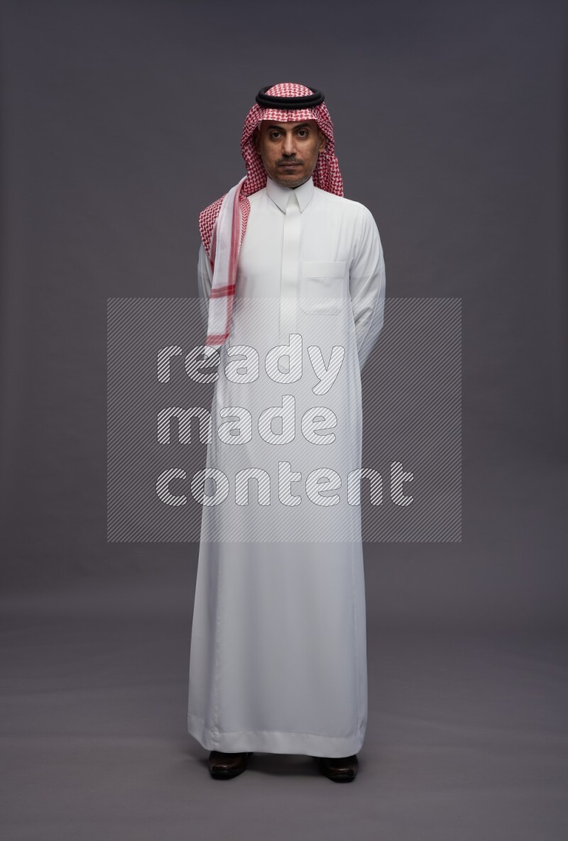 Saudi man wearing thob and shomag standing interacting with the camera on gray background