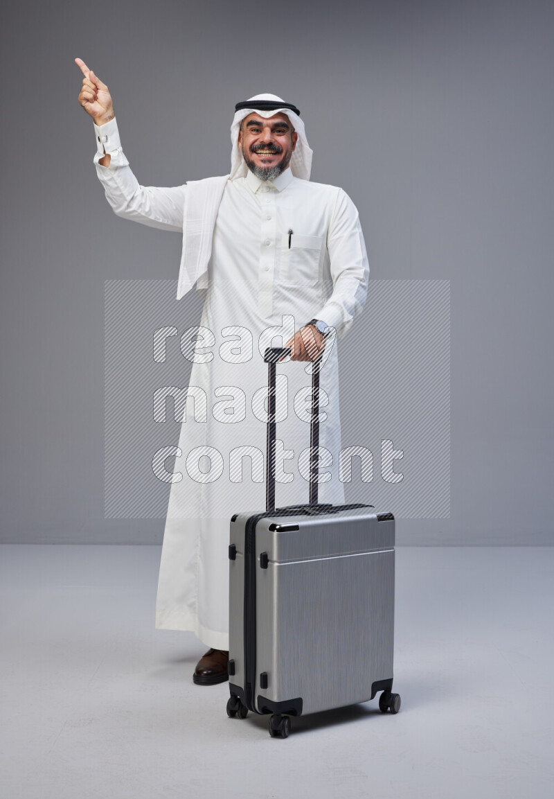 Saudi man wearing Thob and white Shomag standing holding Travel bag on Gray background