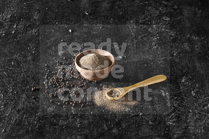 A wooden bowl and a wooden spoon full of black pepper powder with the beads on a textured black flooring