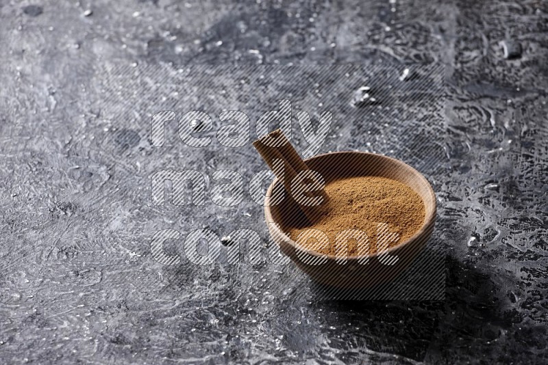 Wooden bowl full of cinnamon powder and a cinnamon stick on a textured black background