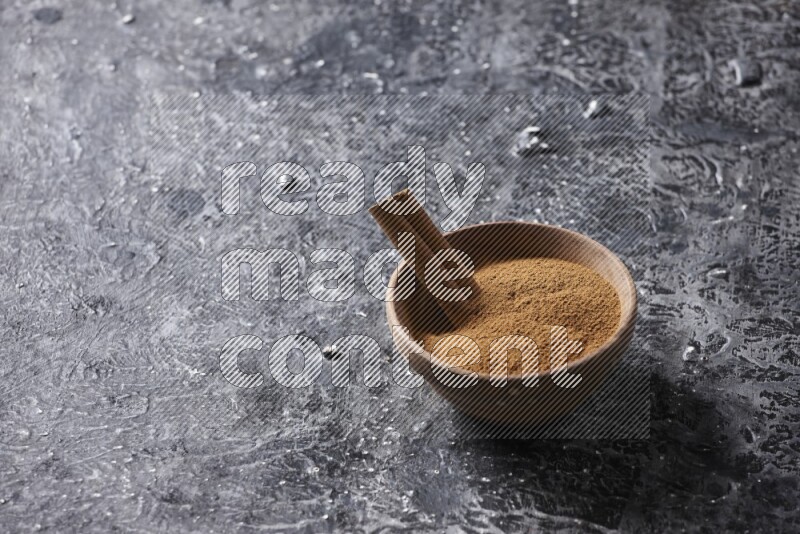 Wooden bowl full of cinnamon powder and a cinnamon stick on a textured black background