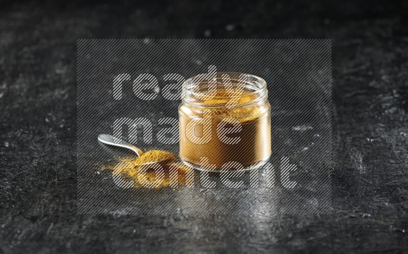 A glass jar and a metal spoon full of turmeric powder on a textured black flooring