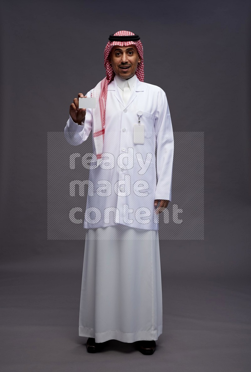 Saudi man wearing thob with lab coat and shomag with pocket employee badge standing holding ATM card on gray background