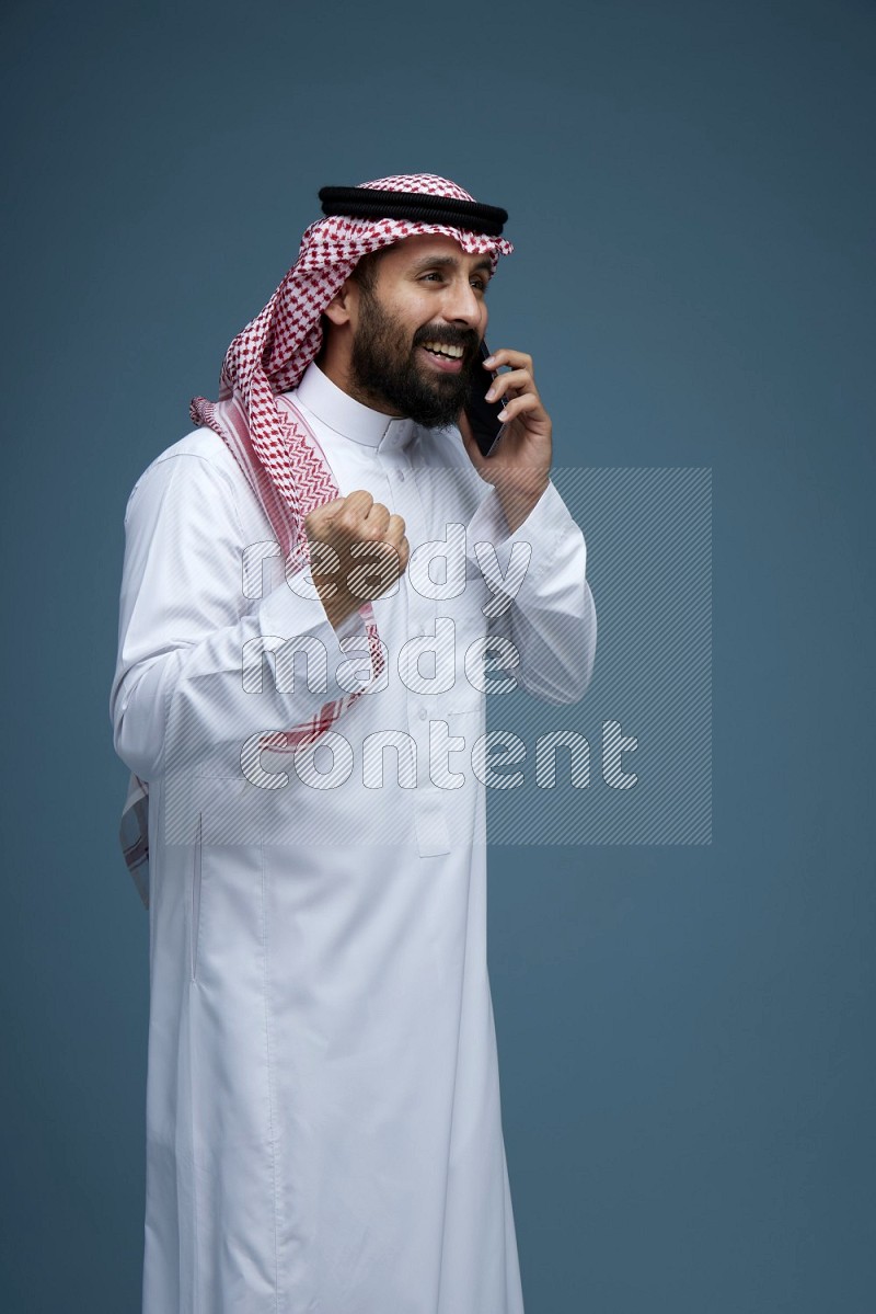 A man having a Call in a blue background wearing Saudi Thob with Shomag