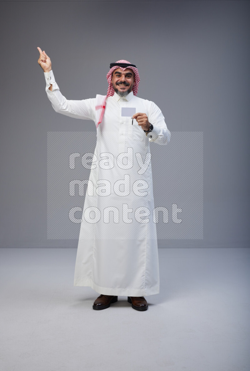 Saudi man Wearing Thob and red Shomag standing holding ATM card on Gray background