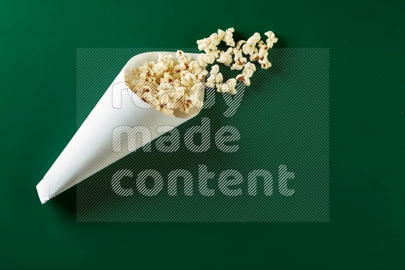 A paper cone full of popcorn and some popcorn came out of it on a green background in a top veiw shot