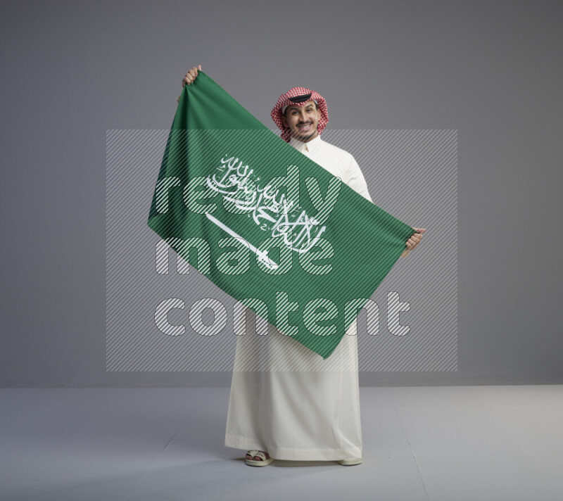 A saudi man standing wearing thob and red shomag holding big saudi flag on gray background