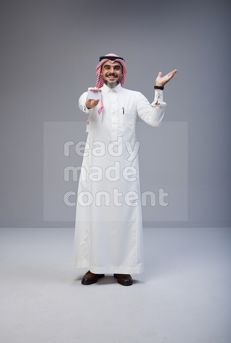 Saudi man Wearing Thob and red Shomag standing holding ATM card on Gray background